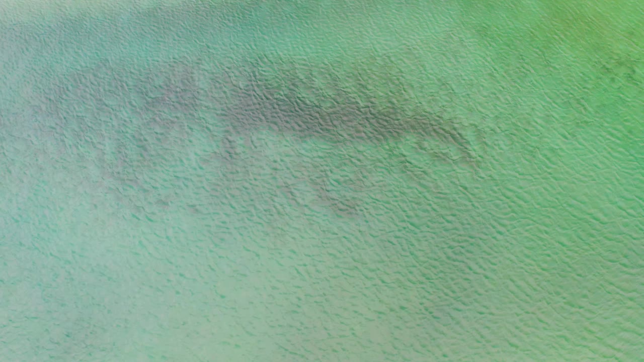 Aerial frame over La Ribera water showing a pale green surface, small wave texture, darker streaking near the upper area, and no visible people.