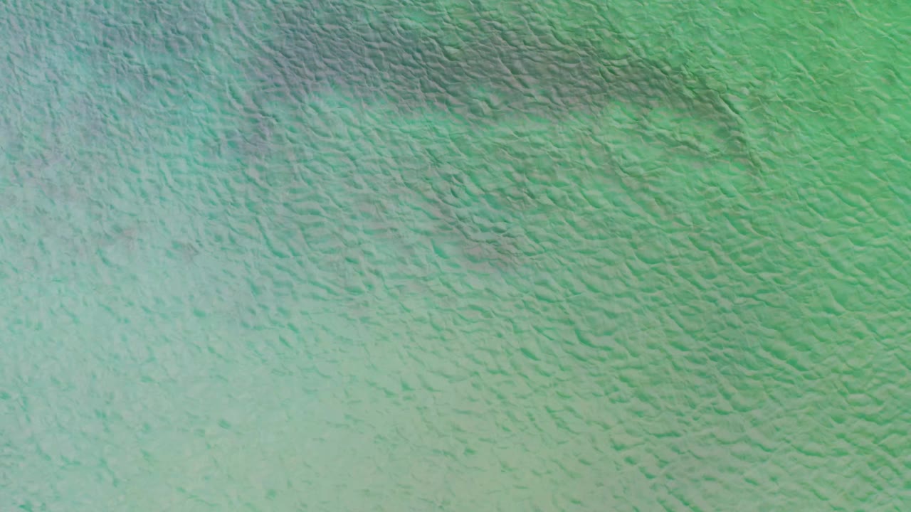 Aerial frame of green-blue saltwater in La Ribera, Baja California Sur, where darker ripple bands spread across the upper water surface with no visible people.