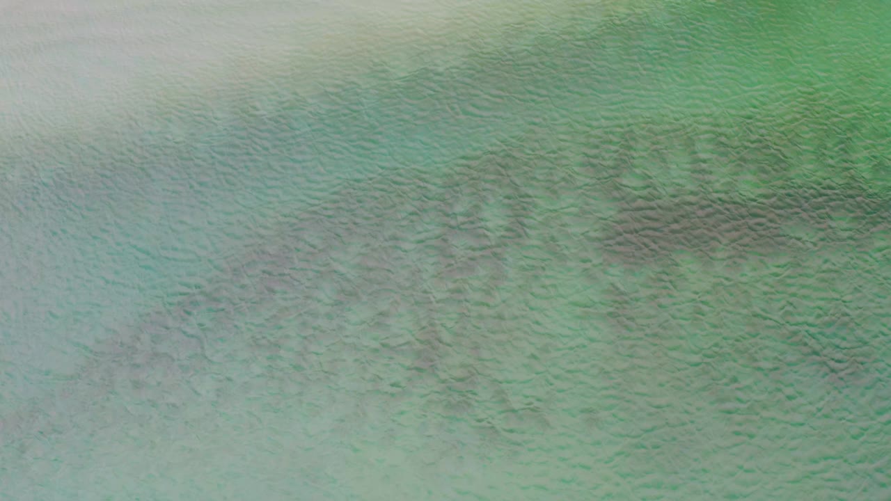 Aerial frame over La Ribera ocean water, showing pale reflected light across the upper left and darker reef-like underwater texture through the center, with no visible people.