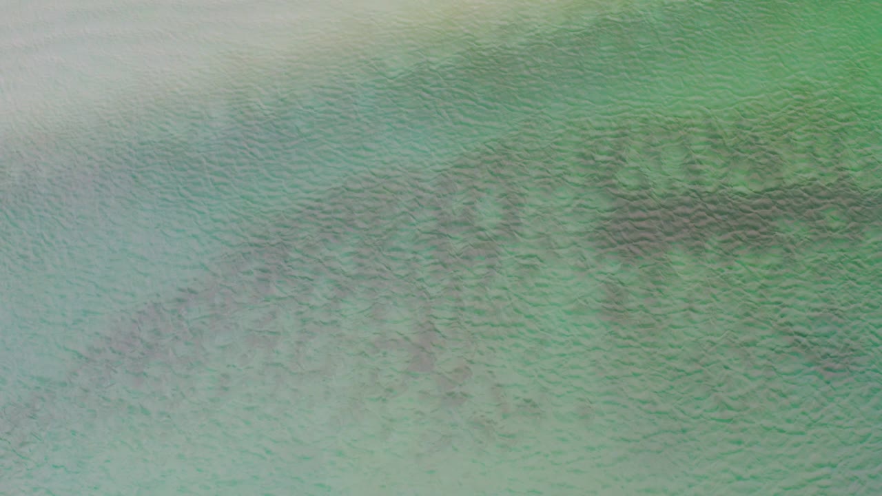 Top-down view of La Ribera coastal water with green tones, fine surface ripples, and muted darker areas below the surface, with no visible people.