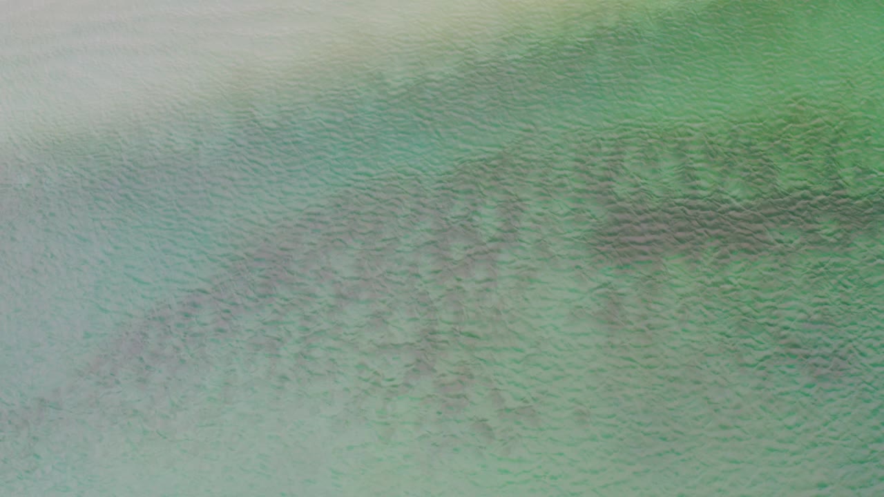 Overhead La Ribera ocean frame with a pale light band crossing the upper left, turquoise water below, and no visible people or vessels.