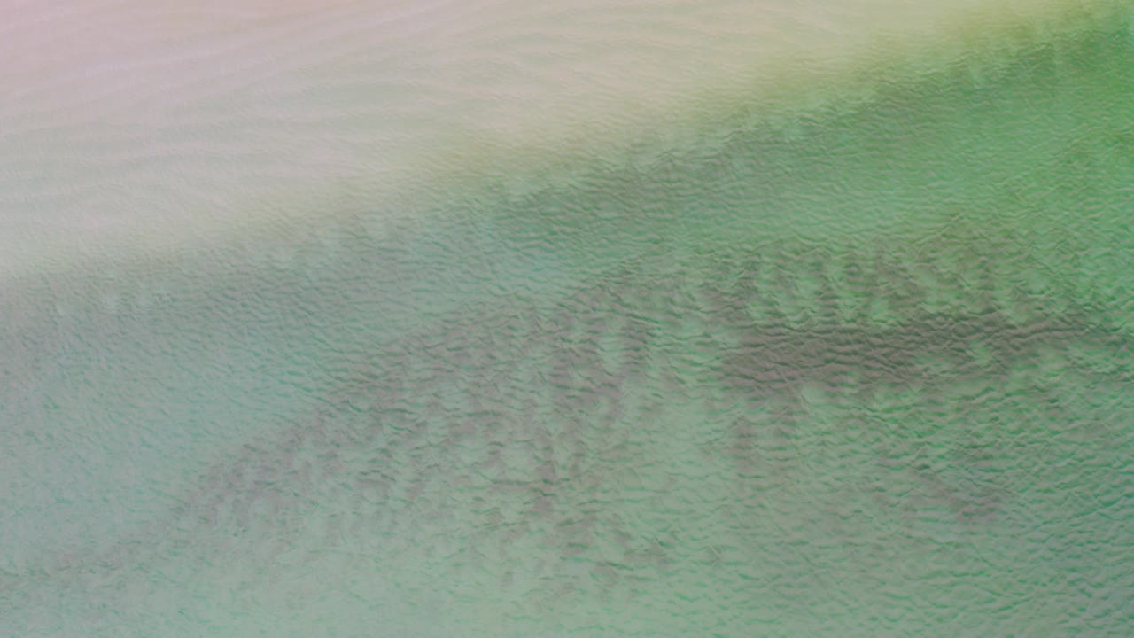 Overhead abstract view in La Ribera, Mexico, with pale sand at the top, green-blue shallow water below, dark submerged streaks across the center, and no visible people.