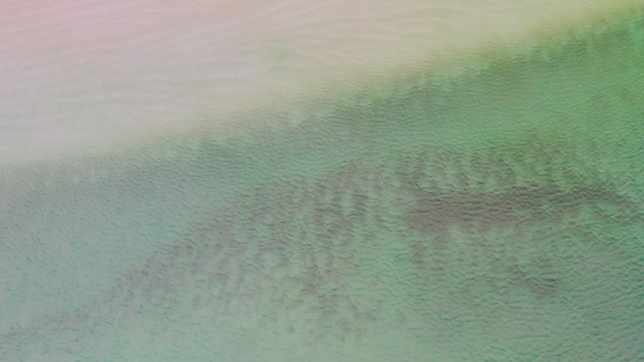 A pastel overhead frame from La Ribera shows a broad pale sand band above turquoise-green water, with a darker submerged pattern slightly right of center and no visible people.