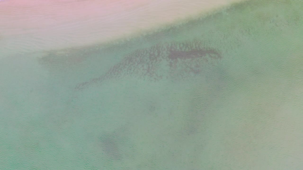 Top-down view of the La Ribera shoreline with pale sand across the top, green-blue shallow water below, faint dark underwater patches, and no visible people.