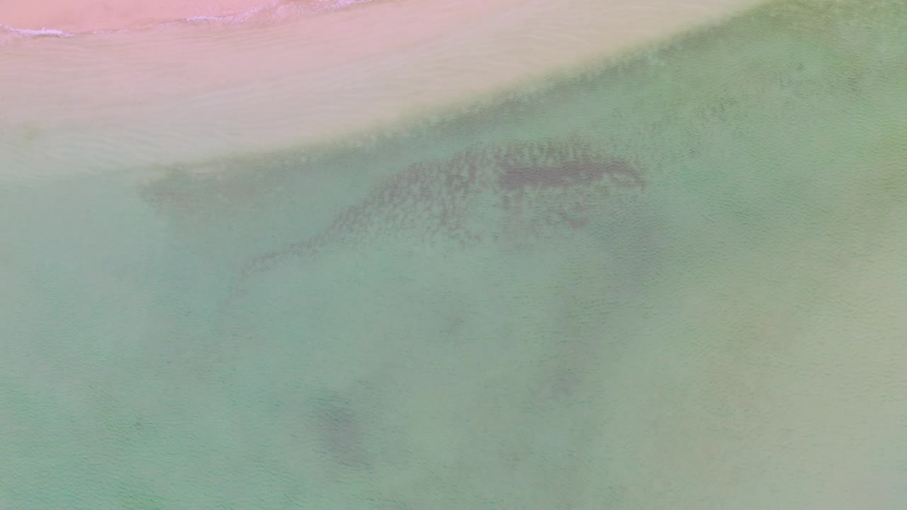 A top-down La Ribera beach view places a thin strip of sand near the top while muted aqua water and shadowy underwater patches spread through the frame, no visible people.