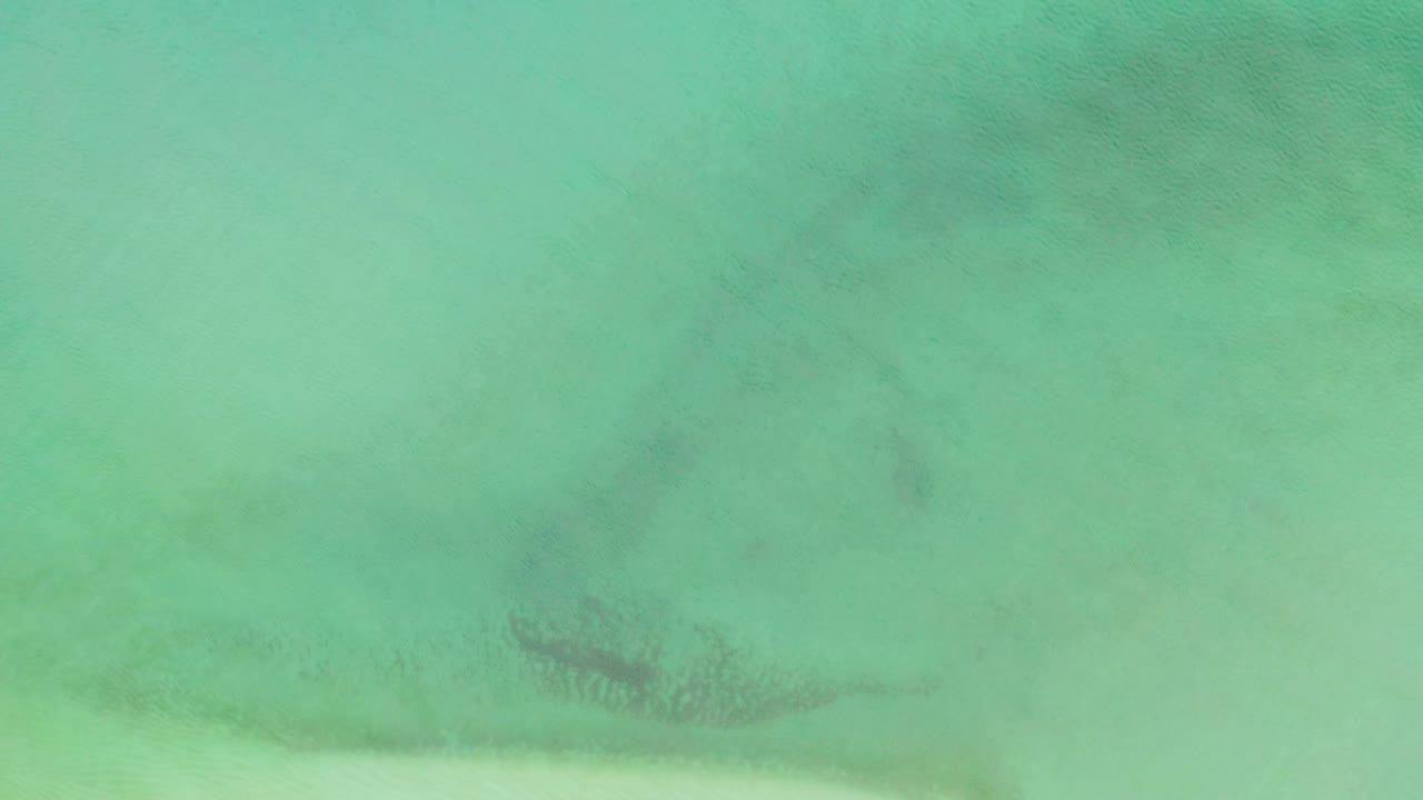 Bird's-eye view of turquoise-green water in La Ribera with pale seabed areas, small darker underwater patches, soft natural light, and no visible people.