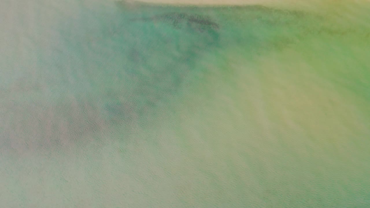 Drone-style overhead frame in La Ribera, Baja California Sur where a pink sand strip begins to read more clearly above green shallow water, with no visible people.