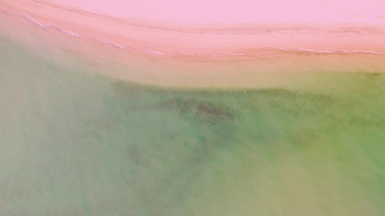 Top-down aerial frame of La Ribera shoreline with pink-toned beach across the top, green shallow water below, darker underwater patches, and no visible people.