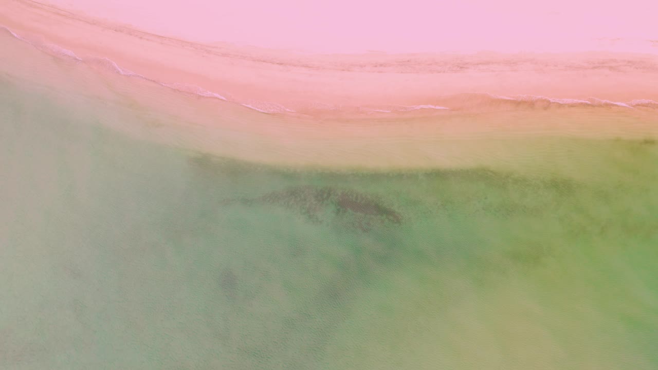 Top-down La Ribera shoreline frame with a broad pink sand area, thin foamy surf, muted green shallows, and no visible people or structures.