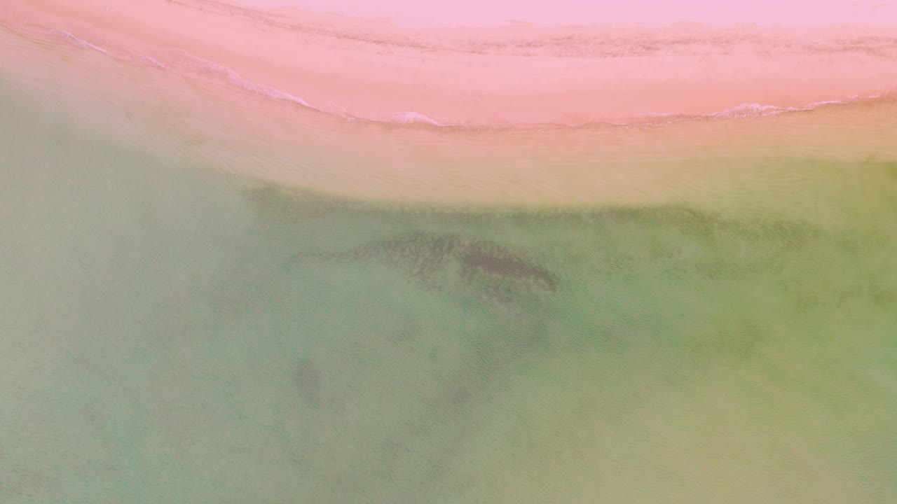 Top-down view of La Ribera shoreline with a pink sand band, pale green shallow water, faint surf line, dark submerged patches, and no visible people.