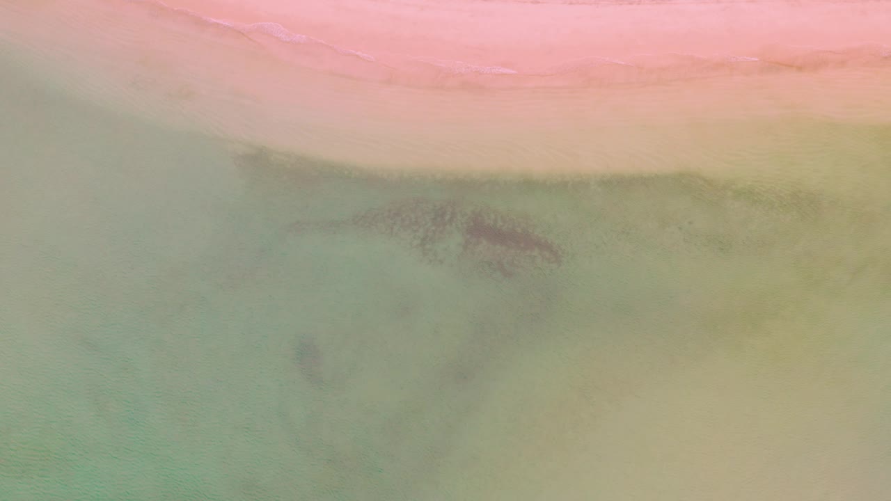 Top-down coastal texture in La Ribera with soft pink shore at the top, muted green water across most of the frame, and a dark central patch beneath the water, no visible people.