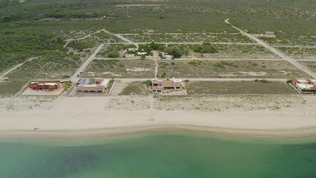 Aerial frame over La Ribera, Baja California Sur showing green-blue seawater, pale sand, four beachside buildings, dirt roads, and scrubland with no visible people.