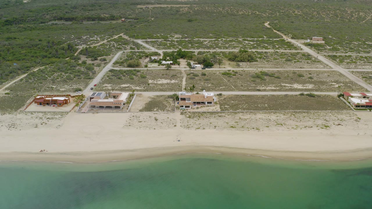 La Ribera aerial frame showing shallow green water, a smooth sandy beach, a row of large beachside structures, pale access roads, and low inland brush with no visible people.