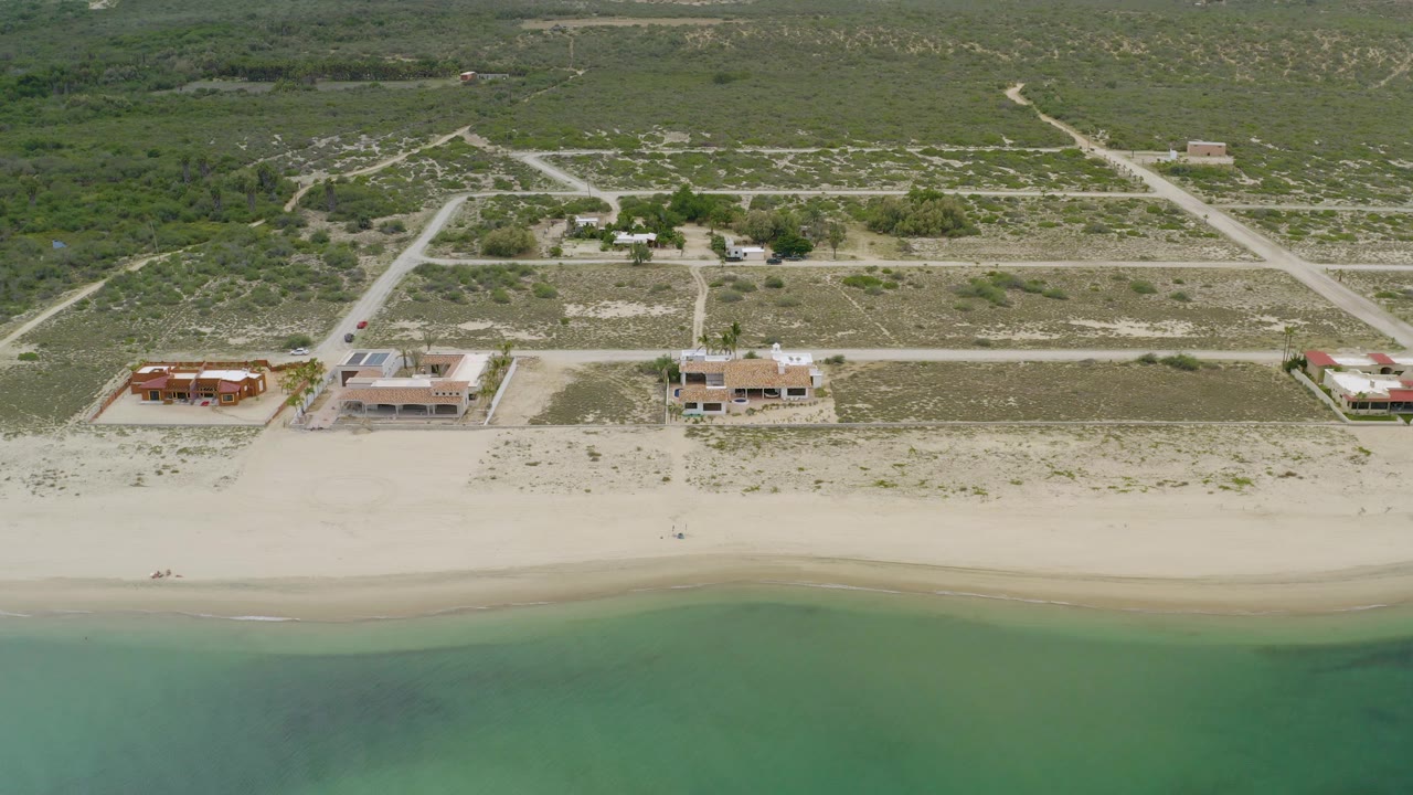 Drone-like coastal view of sand, water, beach buildings, and rural roads.