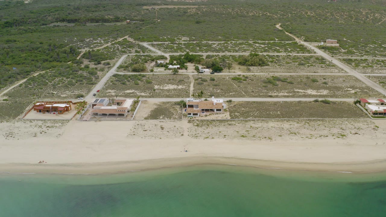 Aerial frame over La Ribera, Baja California Sur showing three beachside houses, pale sand, shallow green-blue water, scrubland, dirt roads, and no visible people.
