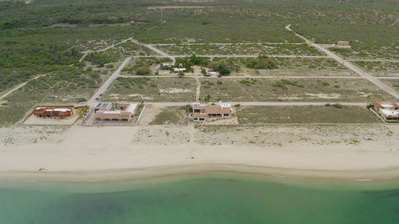 High aerial frame over La Ribera, Baja California Sur showing beach houses in the midground, a smooth sandy beach below, scrubland above, intersecting dirt roads, and no visible people.