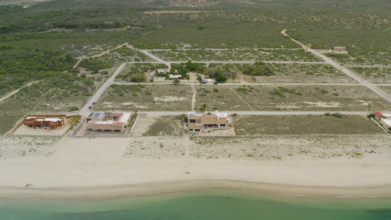 Aerial frame over La Ribera, Baja California Sur showing pale beach, green-blue shoreline, three beachside homes, scrubland roads, and no visible people.