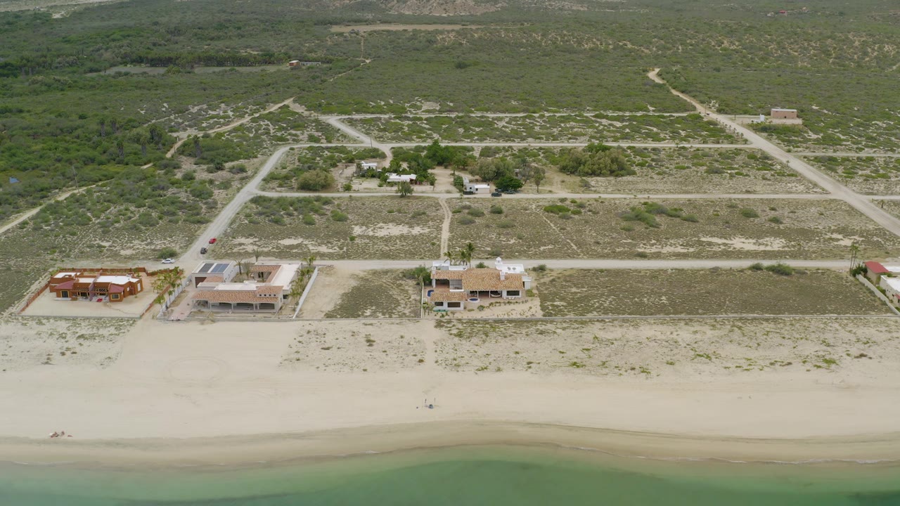 Aerial view of La Ribera, Baja California Sur showing a broad pale beach, several coastal homes with palms, rectangular open lots, dirt tracks, and no visible people.