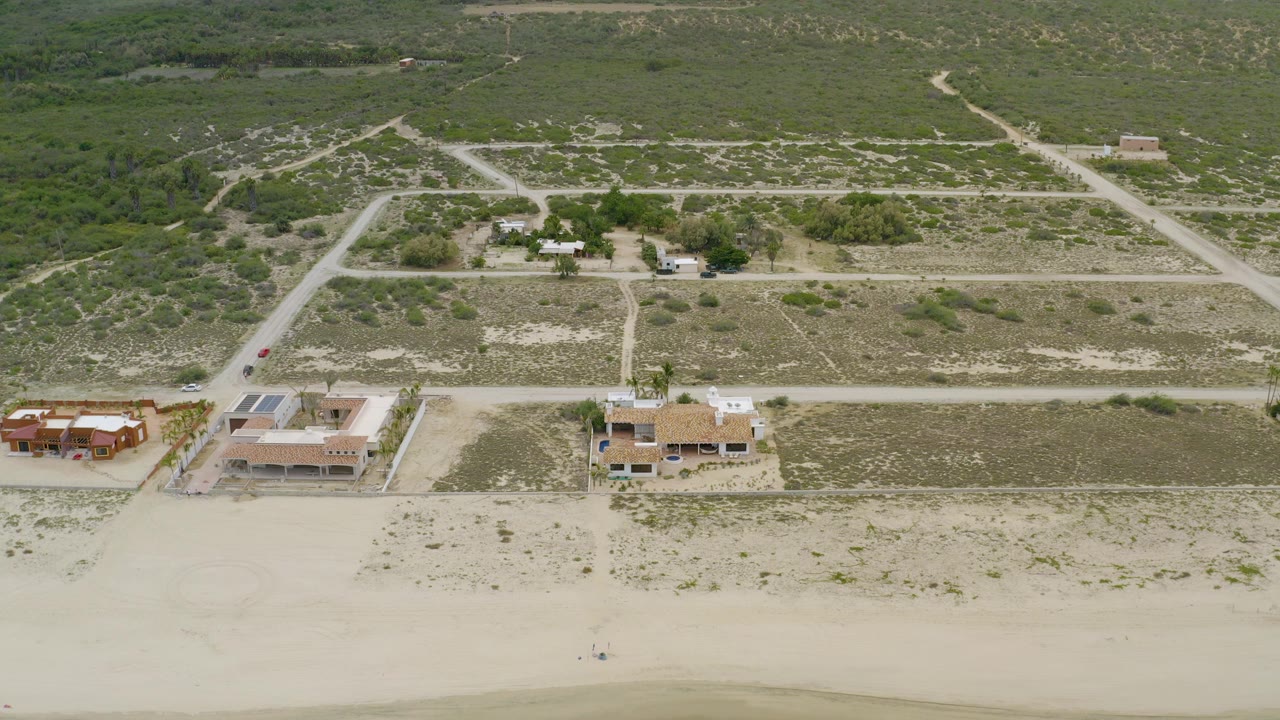 Aerial view in La Ribera, Baja California Sur, with pale beach sand at the bottom of frame, several beachfront houses, dirt roads, inland scrub vegetation, and no visible people.