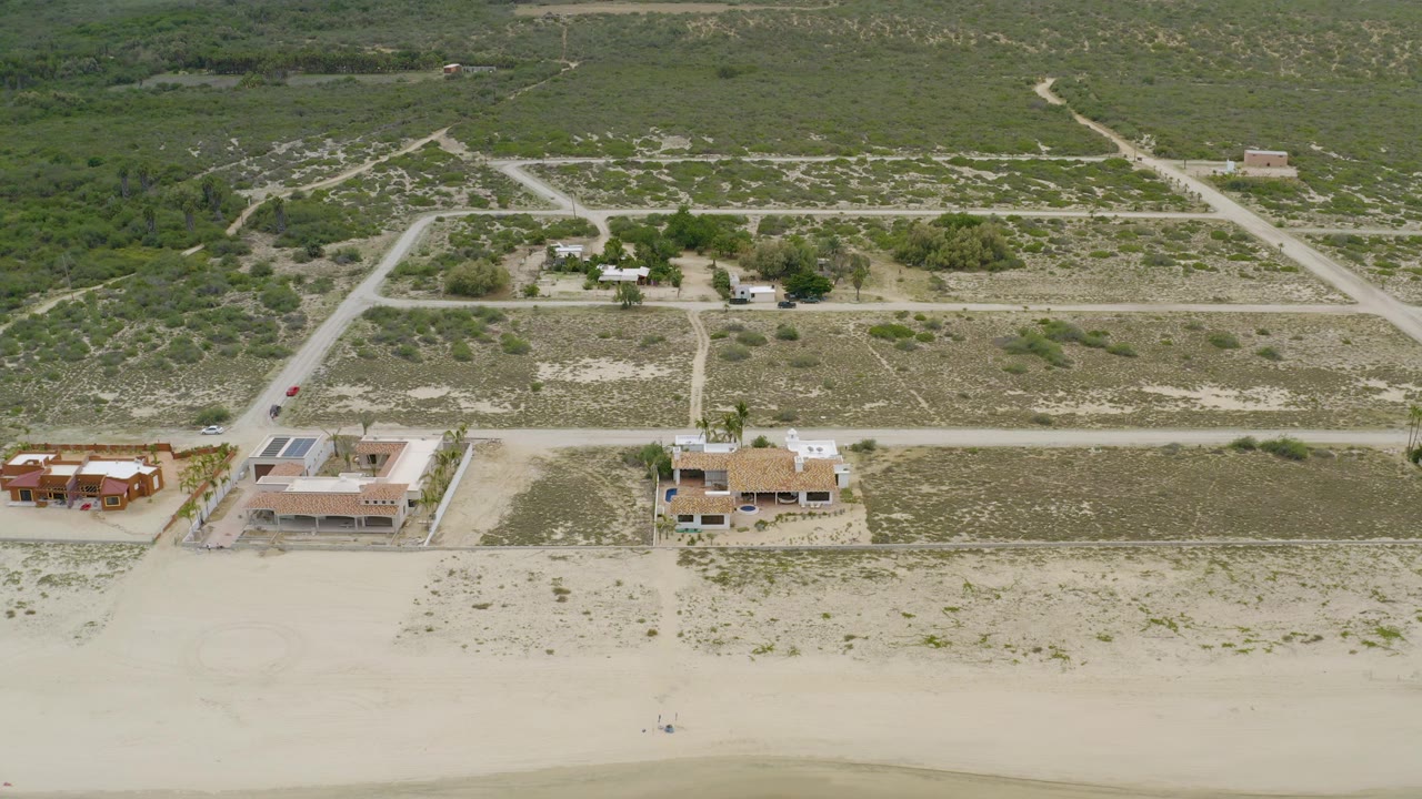 Second aerial frame in La Ribera, Baja California Sur, showing beach sand below, a row of houses near the shore, intersecting dirt roads, green scrubland beyond, and no visible people.