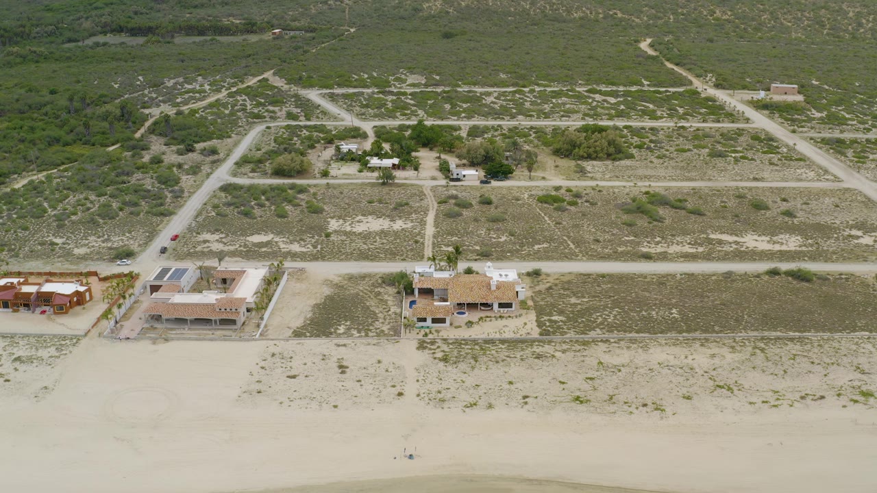 High aerial frame from La Ribera, Baja California Sur, with a sandy beach foreground, separated beachfront homes, a pale dirt-road grid, dense green brush inland, and no visible people.