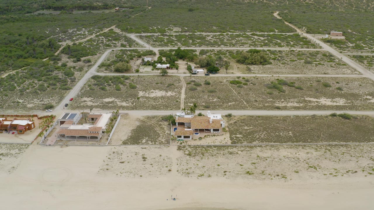 Drone-like view over beachfront homes, empty sand, dirt roads, and low vegetation.