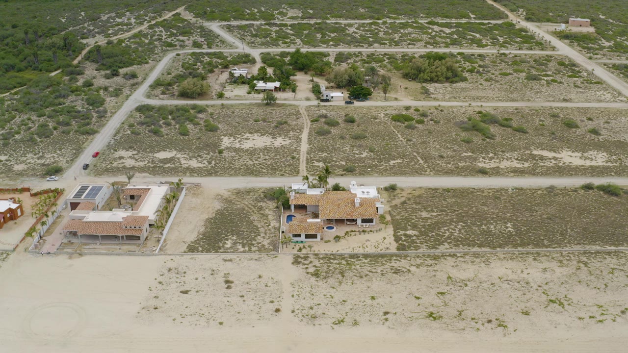 Aerial frame in La Ribera, Baja California Sur, showing two homes near sandy foreground lots, pale roads, scrub vegetation, parked vehicles, and no visible people.