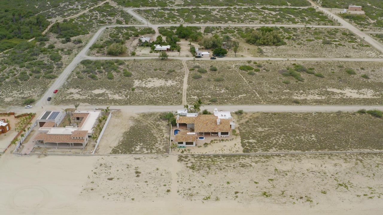Aerial frame from La Ribera, Baja California Sur, showing tile-roof homes, pale unpaved roads, sandy lots, green scrub, a small vehicle on the road, and no visible people.