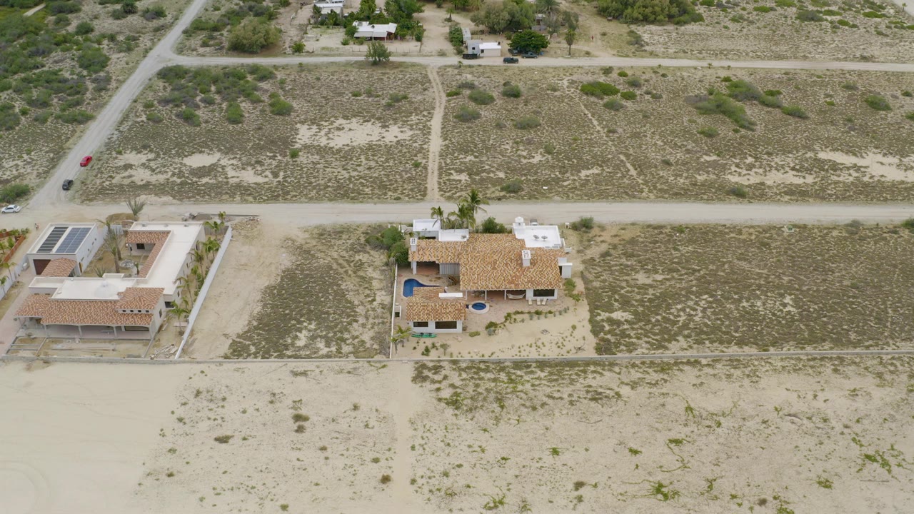 High aerial view in La Ribera, Baja California Sur showing two houses, pale open lots, scrub vegetation, and dirt roads with no visible people.