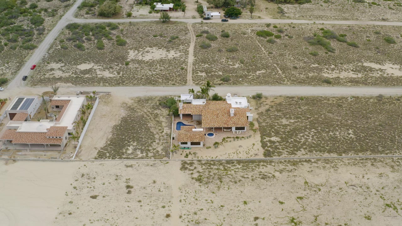 High drone-style view in La Ribera, Baja California Sur showing two private houses, rectangular lot boundaries, dry vegetation, and pale sandy roads with no visible people.