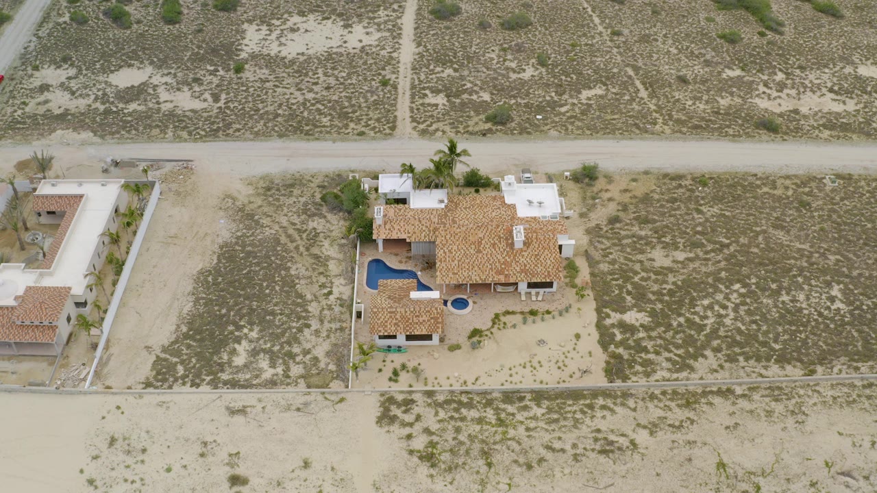 High overhead aerial frame in La Ribera, Baja California Sur, showing a large tan-roofed house, blue curved pool, sandy parcel lines, dirt road, palms, and no visible people.