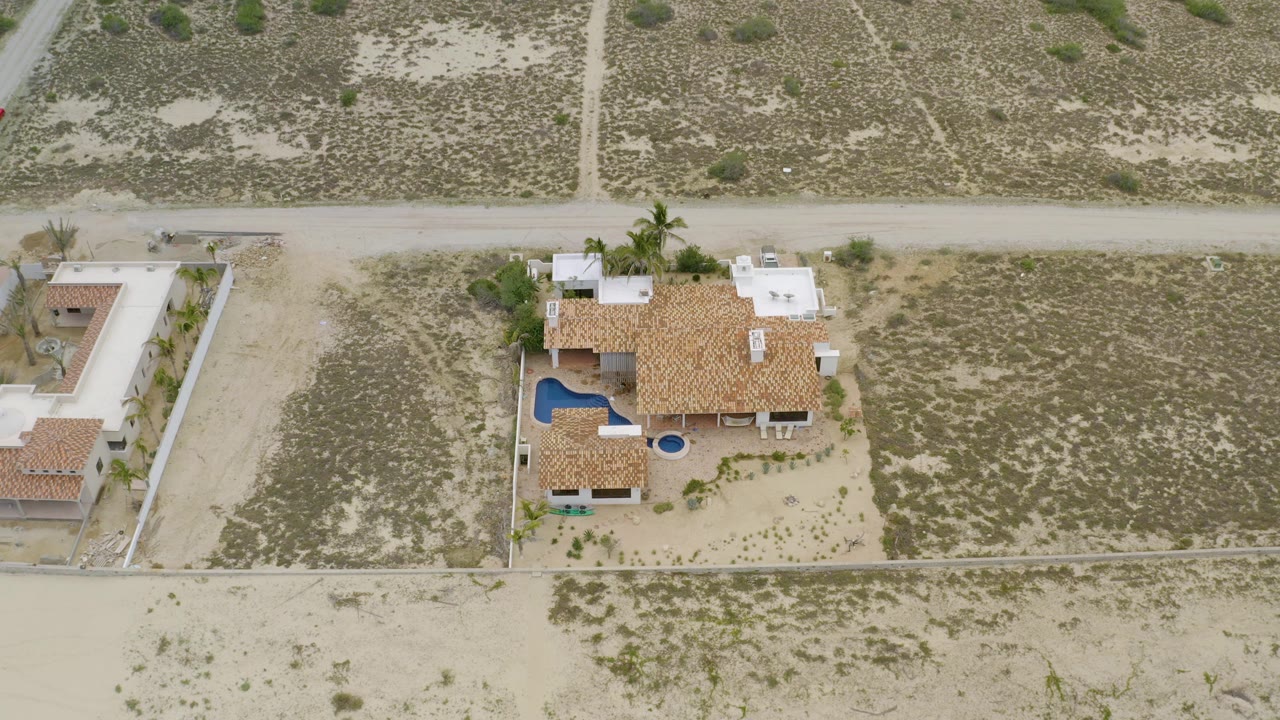 Drone frame over La Ribera, Baja California Sur, with the tan-roofed residence near center, blue pool on the left side of the property, open scrub lots around it, and no visible people.