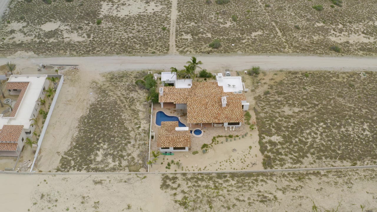 High aerial view in La Ribera, Baja California Sur, showing the walled home site, tile roof, blue pool, neighboring sandy lots, dirt road network, and no visible people.