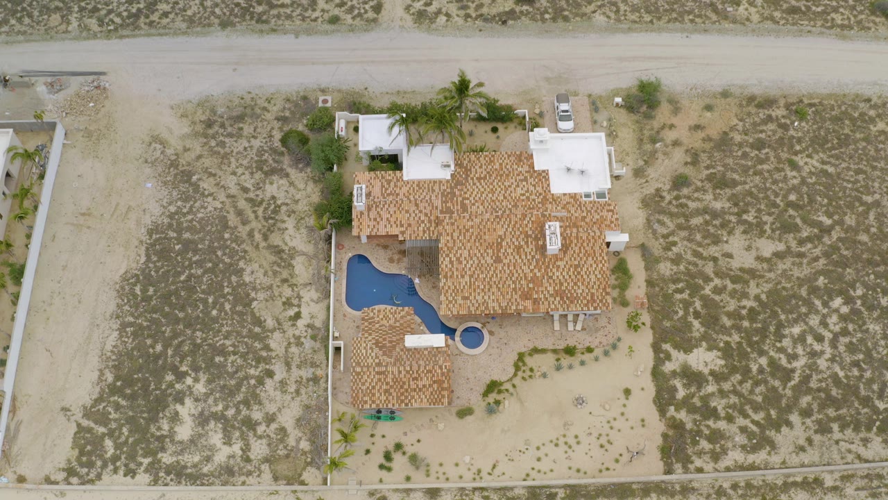 Overhead frame in La Ribera, Baja California Sur, showing a red tile roof home, blue pool, palm trees, sandy roads, scrub lots, and no visible people.