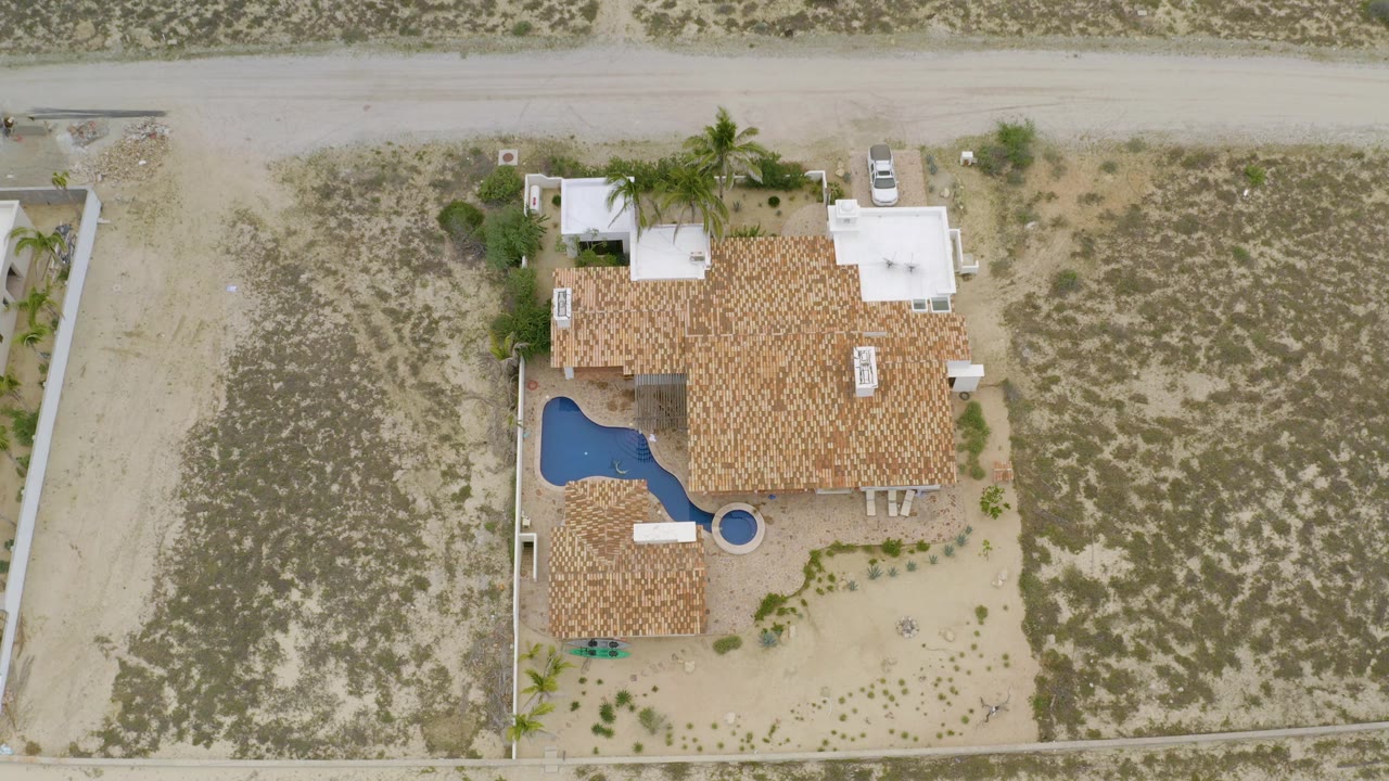 Top-down La Ribera frame with the house roof near center, a blue pool on the left side of the property, a white car by the road, dry lots, and no visible people.