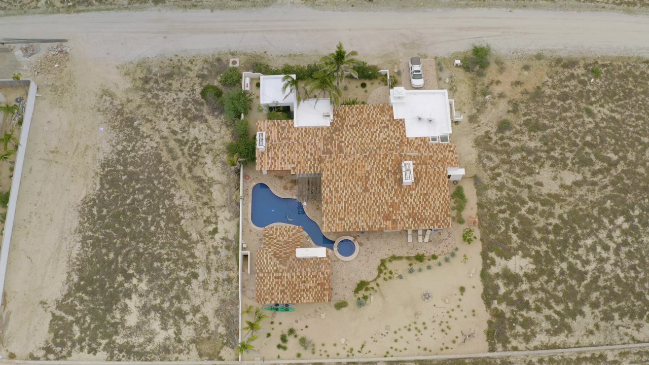 Aerial view in La Ribera with the red roof house slightly right of center, pool at the left edge of the home, sandy road above, scrub lot to the side, and no visible people.