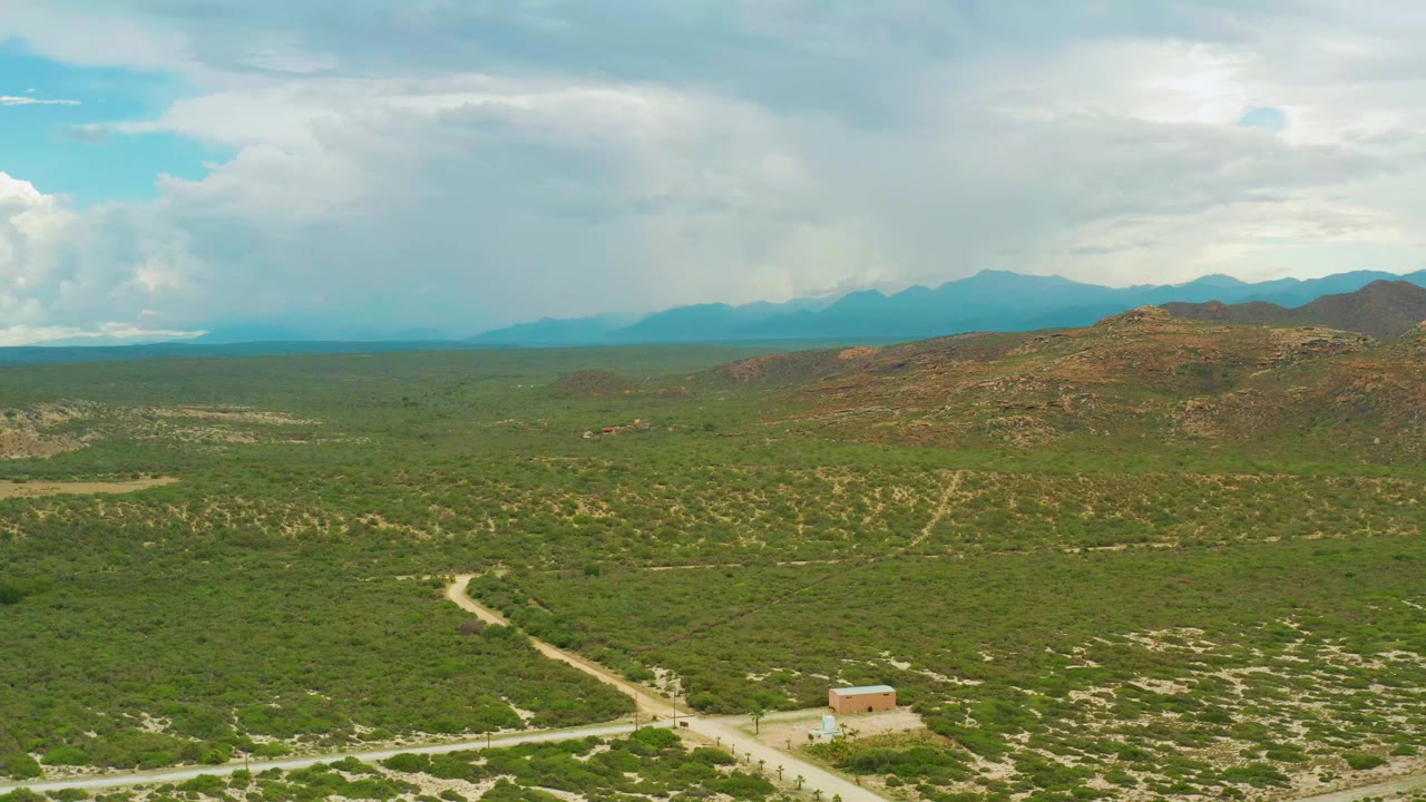 Opening frame over La Ribera, Baja California Sur, with green scrubland, pale dirt roads, a small building near the bottom edge, distant mountains, broad clouds, and no visible people.
