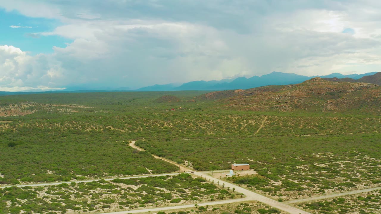 Opening aerial frame over La Ribera, Baja California Sur, with green scrubland, intersecting dirt roads, a small tan house near the lower center, distant mountains, and no visible people.