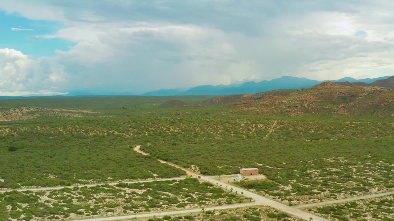 Wide aerial frame from La Ribera, Baja California Sur, with the small house near the lower center-right, pale roads cutting through green scrub, low hills, hazy mountains, and no visible people.