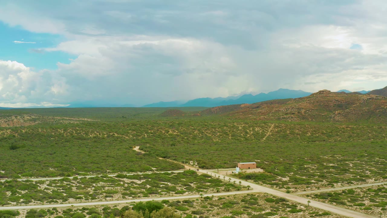 La Ribera landscape frame with the road intersection more centered, green bushes across the plain, a small tan building below the hills, distant blue mountains, and no visible people.