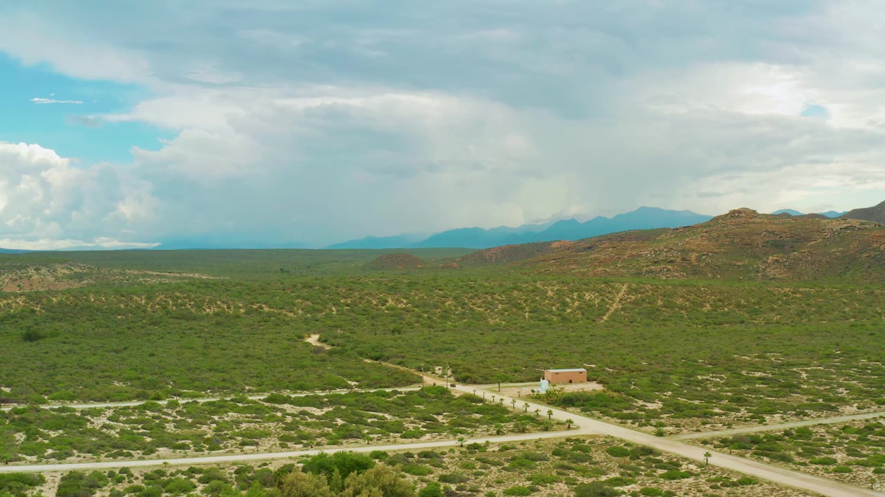 A wide La Ribera frame shows scrub-covered land, pale dirt tracks, the tan building near the lower center, brown hills to the right, a hazy mountain ridge, and no visible people.