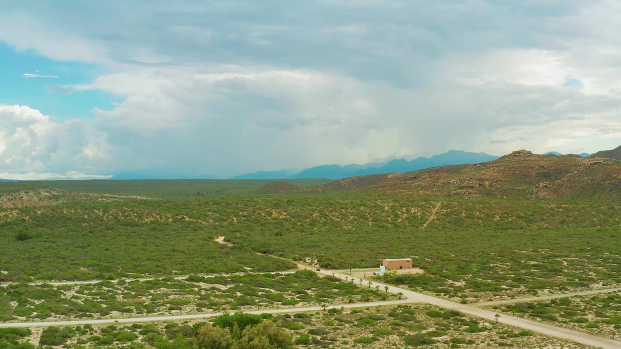 Open green plain, winding pale road, small tan building, hills, mountains, and cloud bank.