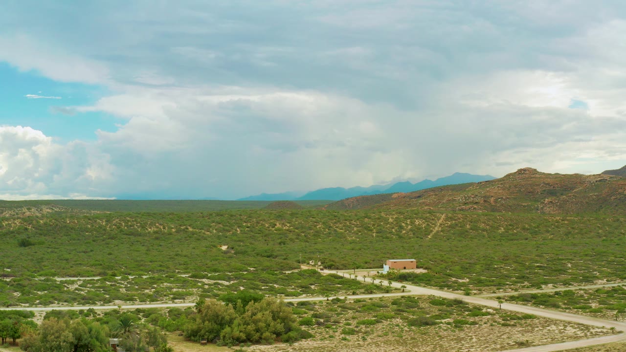 Opening aerial frame over rural La Ribera, Baja California Sur, with green scrubland, pale farm roads, a small rectangular building, distant hills, and no visible people.