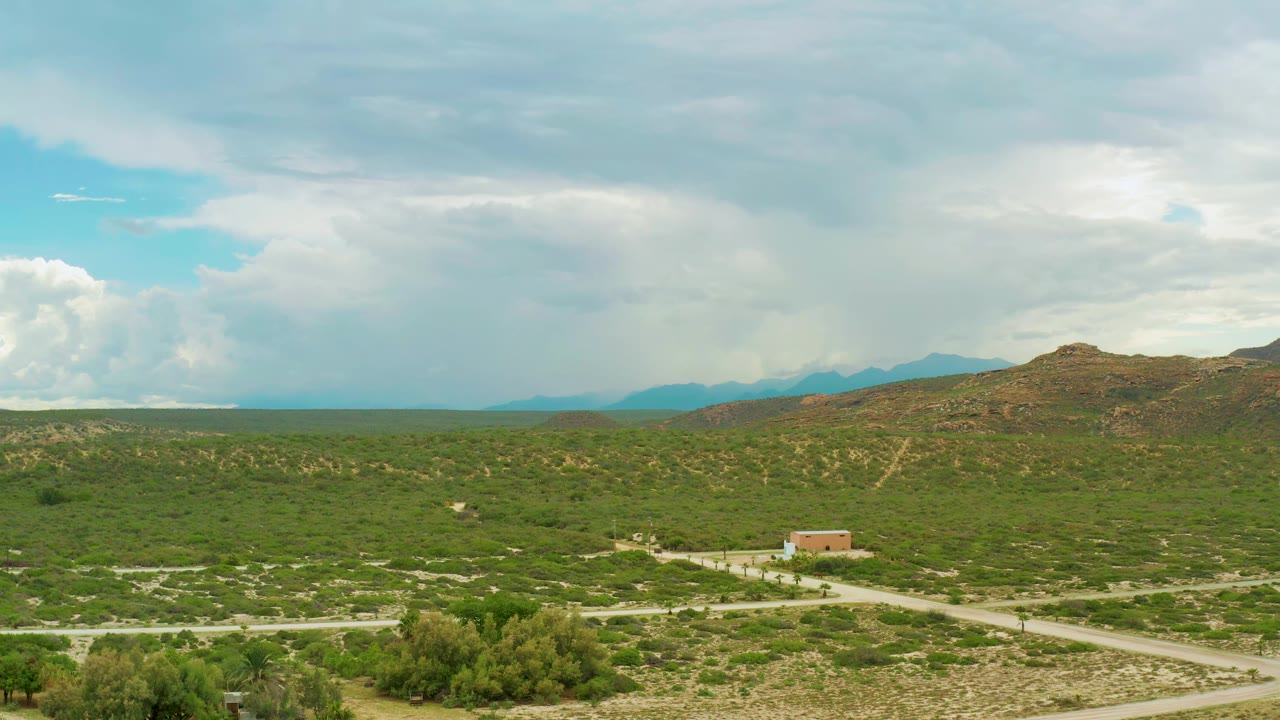Drone view over La Ribera countryside at 0:01, showing a small building near intersecting roads, low green vegetation, brown hills, distant mountains, and no visible people.