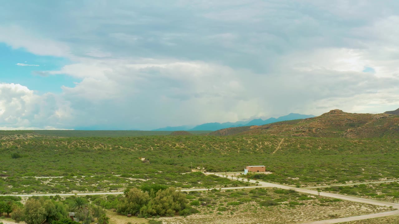 Aerial frame from La Ribera, Baja California Sur, with scrub-covered fields, pale winding roads, a small structure in the midground, hills to the right, and no visible people.