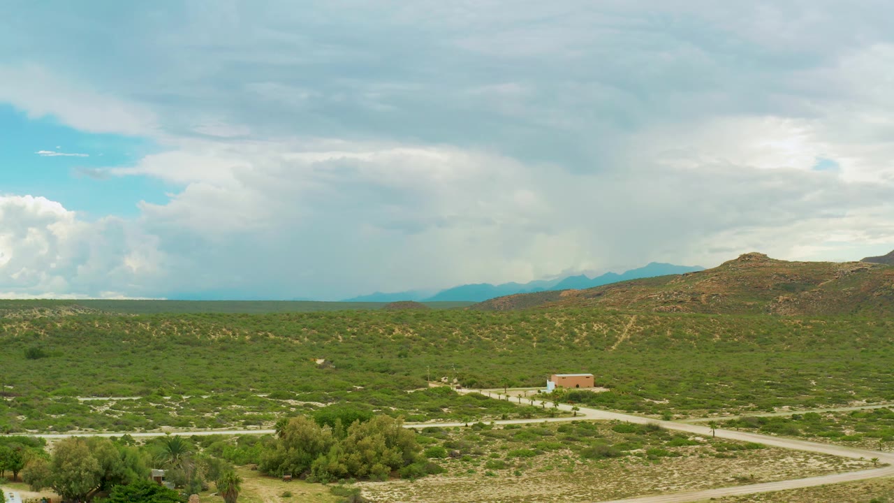 Open green countryside with road crossings, a small building, hills, and cloudy daylight.