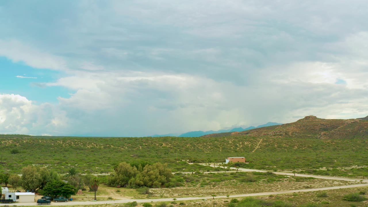 Elevated view in La Ribera with a small tan building below the hill, green scrub across the middle ground, pale dirt roads, a large cloud bank, and no visible people.