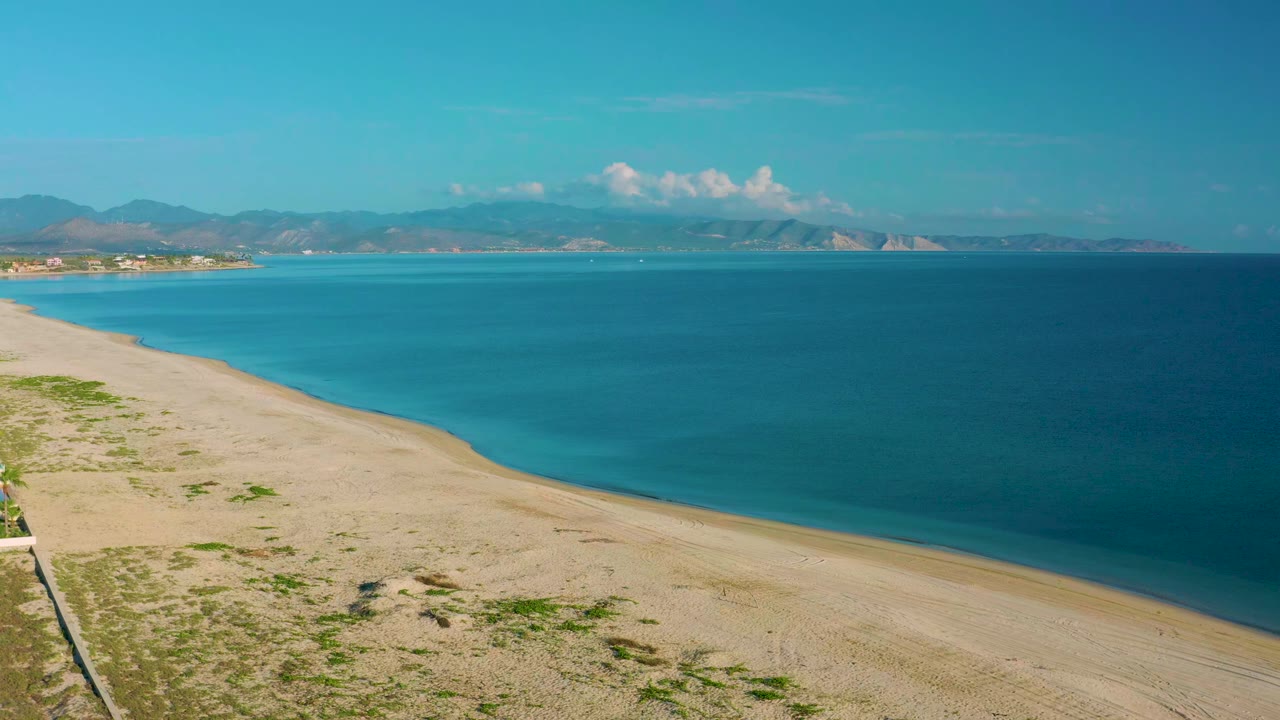 Aerial view over La Ribera, Baja California Sur, with sandy beach in the lower frame, calm blue water, distant mountains, white clouds, and no visible people.