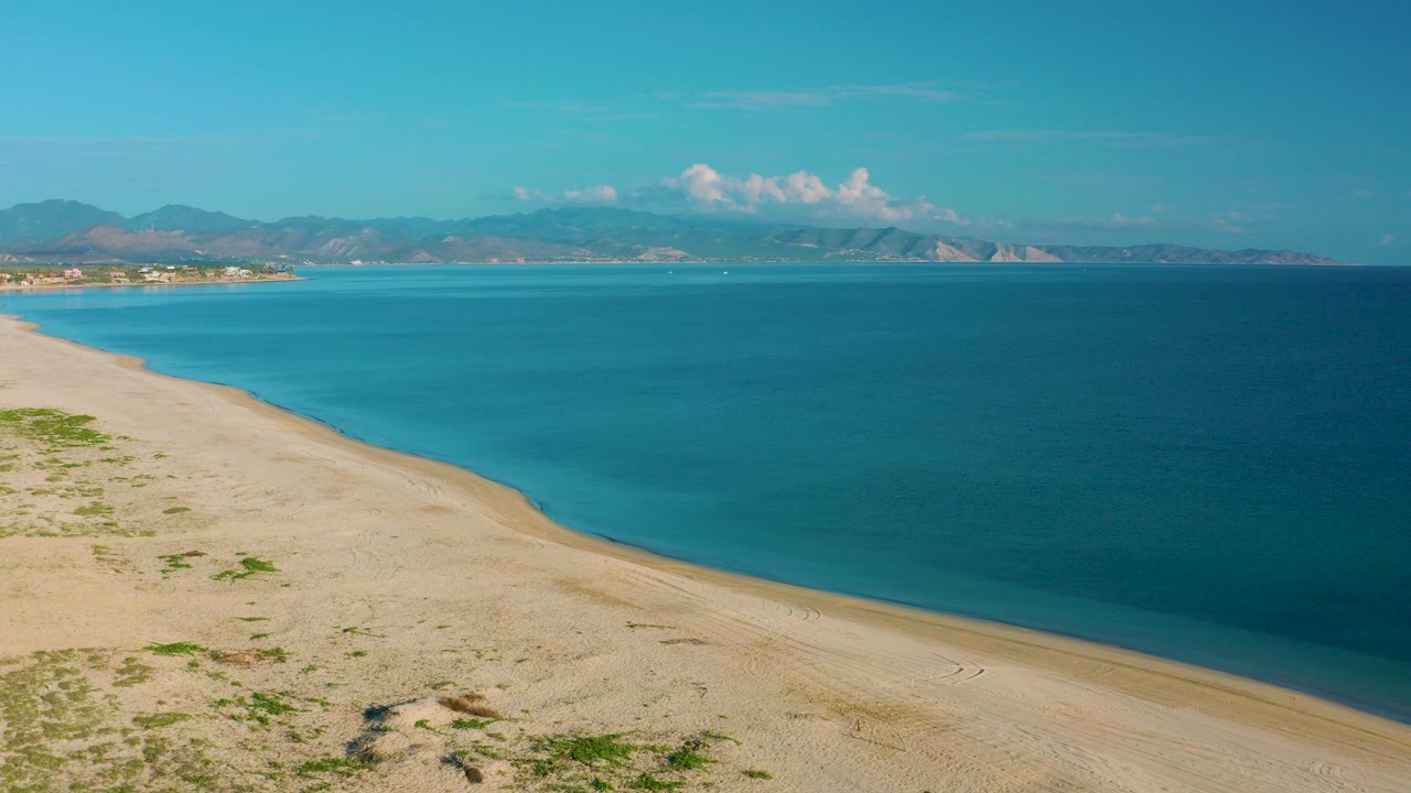 Wide aerial frame of La Ribera, Baja California Sur, with pale sand in the foreground, turquoise water along the shore, distant mountains across the bay, and no visible people.