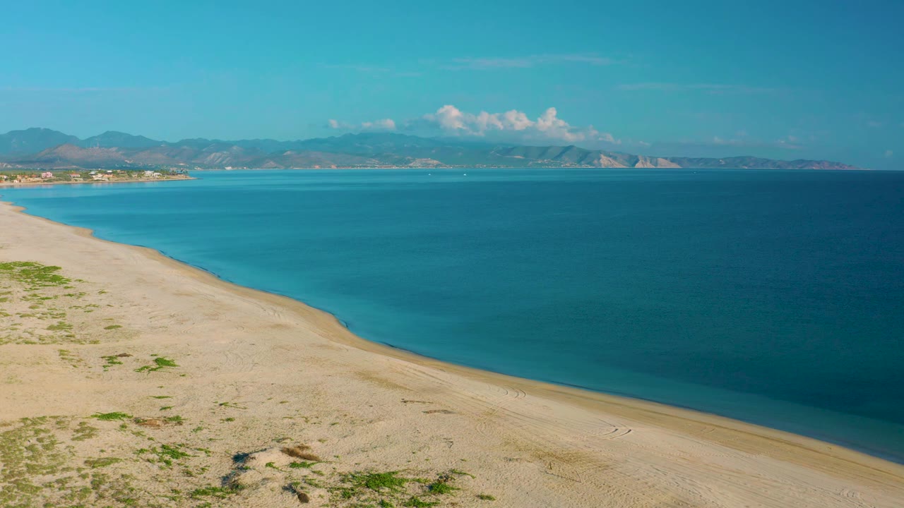 La Ribera shoreline seen from above, with the sandy curve lower left, clear blue water filling the right side, distant mountains on the horizon, and no visible people.